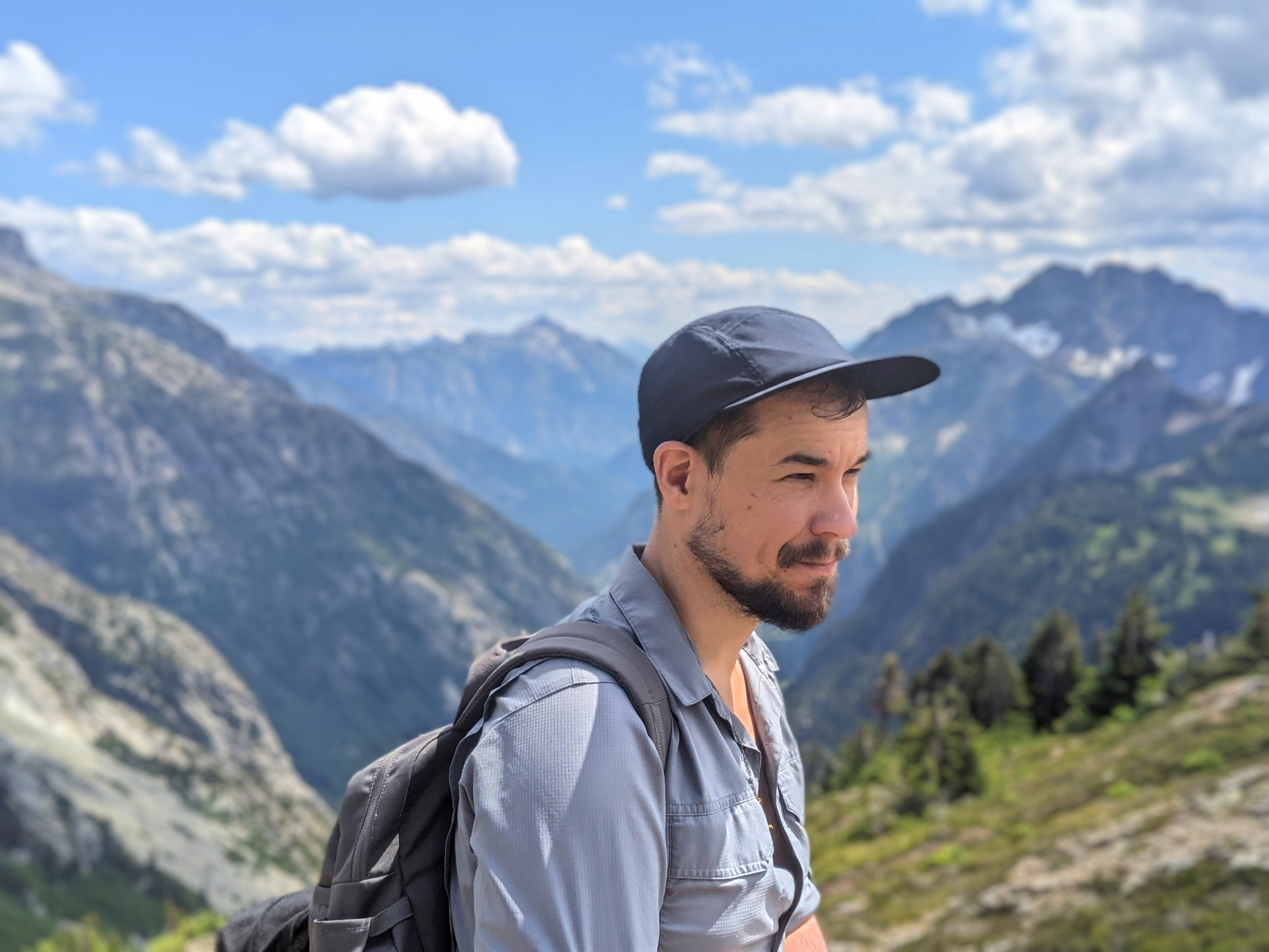 Smiling person wearing ballcap and backpack at altitude with mountains and trees in the background.