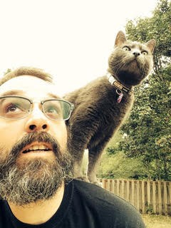 Person with beard and glasses gives side glance with cat standing on and peering over should in same direction.
